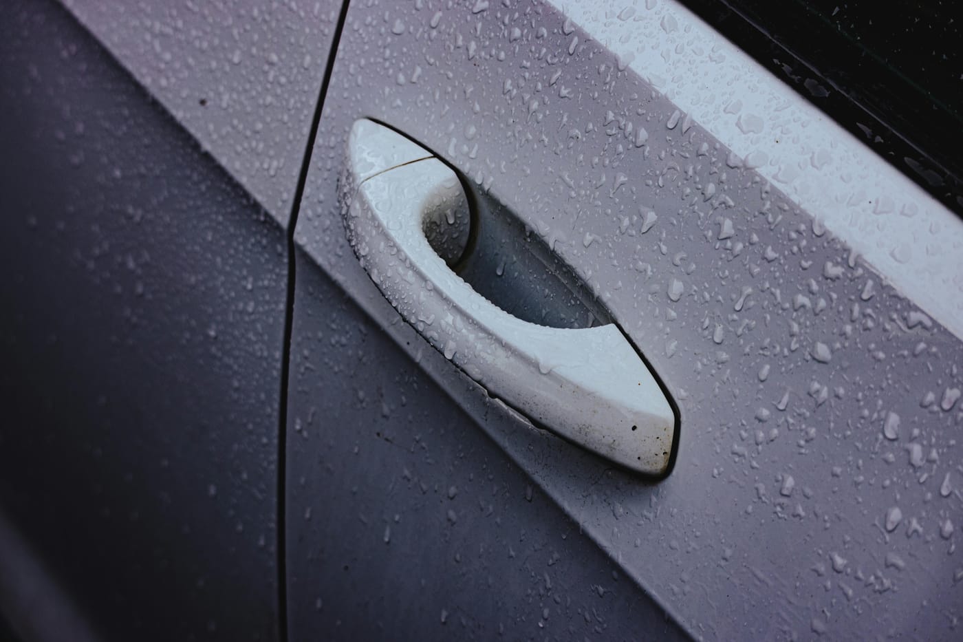 Close-up of a wet silver car door handle covered in rain droplets on a grey vehicle - evoking an emergency lockout in a Washington DC downpour