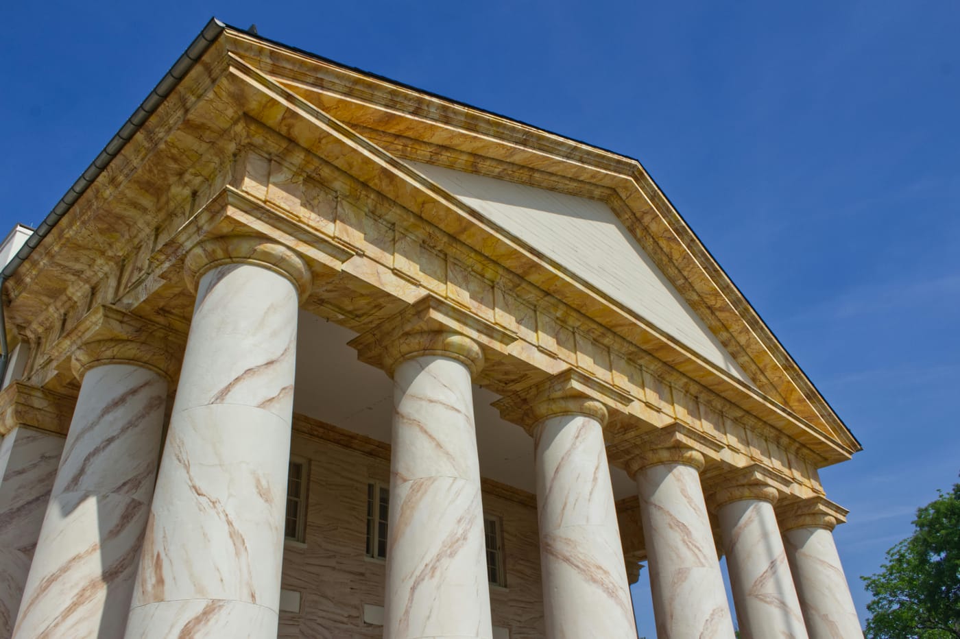 Low-angle view of the stately Arlington House Greek Revival portico with tall marble columns and warm stone pediment against a clear blue sky - evoking the grand neoclassical architecture of Embassy Row