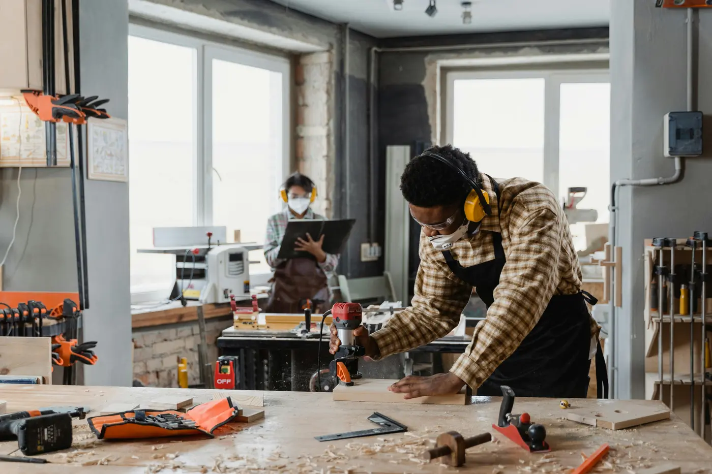 Skilled tradesperson in safety earmuffs and apron operating a power drill at a workshop bench with hand tools, representing the hands-on craft of a locksmith trade career